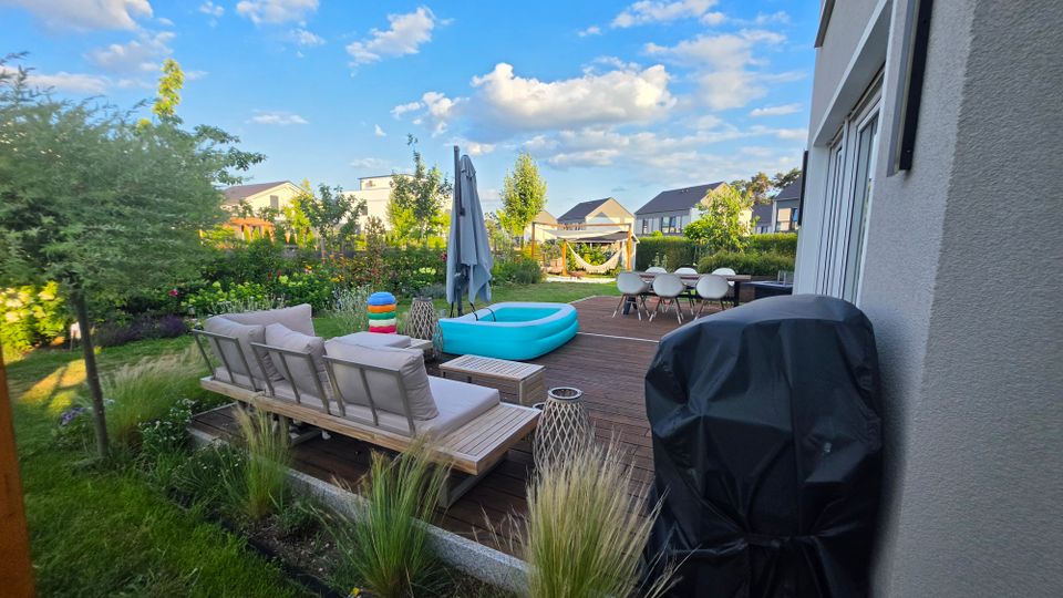 Holzterrasse hinter einem modernen Wohnhaus mit Loungemöbeln, Esstisch, Sonnenschirm und kleinem aufblasbarem Pool, daneben ein abgedeckter Grill. Umgeben von einem bepflanzten Garten mit Hecken und Blick auf benachbarte Häuser unter blauem Himmel mit Wolken.