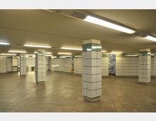 Unterirdischer U-Bahn-Bereich mit weißen, gefliesten Wänden und in regelmäßigen Abständen angeordneten quadratischen Säulen. Der Boden besteht aus grauem Fliesenbelag, die Decke ist mit linearen Leuchtstofflampen beleuchtet, und an einer Wand ist eine Wandgestaltung mit Vogelmotiven zu sehen.