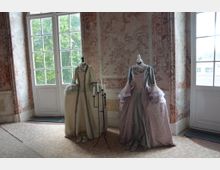 Zwei Schaufensterpuppen in historischen Barockkostümen stehen in einem Raum mit marmorierten Wänden und großen Panoramafenstern, durch die grüne Bäume sichtbar sind. Vor den Puppen befindet sich ein schwarzer Kerzenhalter, und auf dem Boden stehen passende Schuhe.