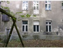 Eine Außenaufnahme eines alten, grauen Gebäudes mit abblätterndem Verputz. Im Vordergrund sind ein schräger Holzpfosten sowie einige Pflanzen und bodennahes Gestrüpp zu sehen, während im Hintergrund mehrere Fenster und eine Tür sichtbar sind.