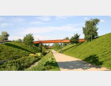 Eine parkähnliche Landschaft mit einem gepflasterten Fußweg, der zwischen zwei grasbewachsenen Erdhügeln verläuft. Eine rostfarbene Stahlbrücke verbindet die Hügel, während Bäume und Sträucher entlang des Weges und der Böschungen stehen.