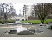 Ein kleiner Park mit einem trockenen Brunnen im Vordergrund, umgeben von gepflasterten Wegen, Rasenflächen und Bäumen. Im Hintergrund führt eine breite Treppe mit Balustraden zu erhöhten Bereichen, daneben ein Wohngebäude.