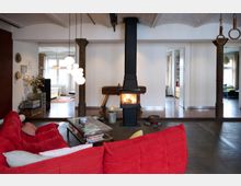 Wohnzimmer in einem offenen, loftartigen Raum mit einem roten Sofa, einem Glastisch und einer schwarzen Kaminheizung in der Mitte. Im Hintergrund sind mehrere Räume mit großen Fenstern, rustikalen Holzsäulen und moderner Einrichtung sichtbar.