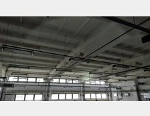 Industriehalle mit hoher Decke aus Beton und rechteckigen Vertiefungen. An der Decke sind lange, hängende Lampen und Rohrleitungen befestigt, die über den Raum verlaufen; an den Wänden befinden sich mehrere Fenster mit Milchglas.