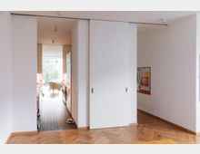 Minimalistisch gestalteter Wohnraum mit Schiebetüren, die einen Durchgang zu einer modernen Küche mit Fliesenboden und einer großen Fensterfront freigeben. Der Raum hat Parkettboden und helle Wände, im Hintergrund sind ein Stuhl und ein Baum außerhalb des Fensters sichtbar.