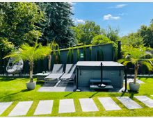 Garten mit Rasenfläche, flankiert von Palmen in Töpfen. Im Vordergrund befindet sich ein gepflasterter Pfad, der zu einer Terrasse mit zwei Sonnenliegen und einem Whirlpool führt. Im Hintergrund stehen hohe Bäume und eine graue Gartenmauer.