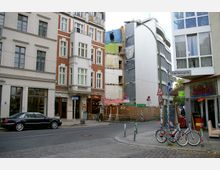 Straßenszene mit mehrstöckigen Gebäuden, darunter ein weißes und ein rot-weißes Wohnhaus, sowie ein modernes Gebäude mit abgerundeten Fassaden rechts. In der Mitte ist ein Gebäude im Umbau sichtbar, daneben stehen Straßenschilder, parkende Autos und Fahrräder sowie ein Café mit roten Sonnenschirmen.