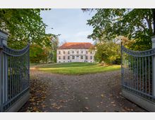 Herrenhaus mit rosa Putzfassade und rotem Satteldach, umgeben von einer parkartigen Anlage mit gepflegter Rasenfläche und großen Bäumen; der Blick führt durch ein geöffnetes, schmiedeeisernes Tor.