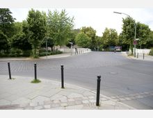 Straßenszene mit einer gepflasterten Kreuzung, umgeben von Bäumen und Grünflächen. Im Hintergrund befindet sich eine kleine Fußgängerbrücke mit Geländern, daneben stehen Straßenlaternen und Poller zur Abgrenzung.