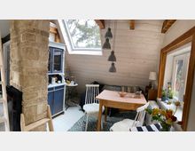 Gemütlicher Wohnraum mit Dachschräge, einem großen Fenster, einer Holzwandverkleidung und sichtbaren Holzbalken. Im Raum stehen ein Holztisch mit Stühlen, ein blauer Vitrinenschrank und eine gemauerte Säule, daneben Pflanzen und Dekorationen auf der Fensterbank.