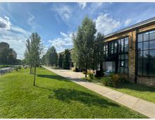 Ein niedriger Industriegebäudekomplex mit großen Glasfenstern und Backsteinfassade, flankiert von einer gepflasterten Gehwegfläche und einer grünen Rasenfläche mit Bäumen, unter einem Himmel mit vereinzelten Wolken.