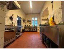 Professionelle Großküche mit Edelstahl-Arbeitsflächen, Gasherd, Waschbecken und Küchengeräten. Der Raum hat weiße Fliesenwände mit kleinen Mustern, einen terrakottafarbenen Boden und ein großes Fenster im Hintergrund.