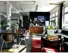 Heller, loftartiger Wohnraum mit offener Küche im Hintergrund, einem langen Holztisch mit Stühlen und einer grünen Sofalandschaft neben einem Glastisch im Vordergrund. Große Fenster lassen viel Tageslicht herein, während zahlreiche Pflanzen das industrielle Design ergänzen.