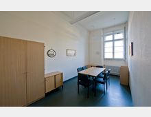 Ein kleiner Besprechungsraum mit einem rechteckigen Holztisch und sechs gepolsterten Stühlen. An der Wand befinden sich ein großer Holzschrank, ein kleiner Schrank, zwei Bilder, eine Uhr und ein großes Fenster mit Sprossen, das Tageslicht hereinlässt.