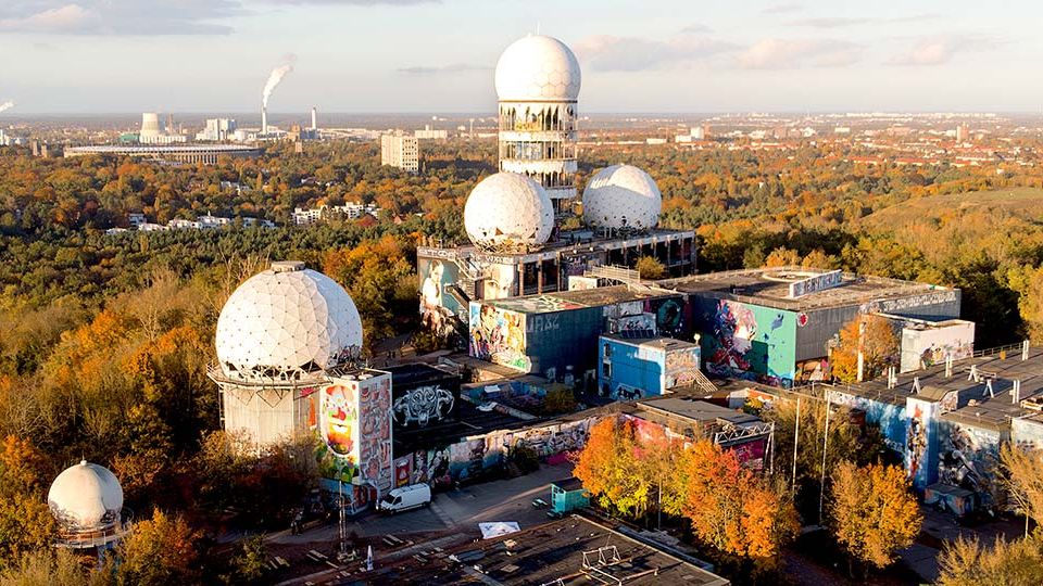 Luftaufnahme einer ehemaligen Radar- und Abhörstation im Wald, mit mehreren großen weißen Radarkuppeln (Radomen) auf flachen, teils verfallenen Gebäuden. Die Anlagen sind mit Graffiti bedeckt und von herbstlich gefärbten Bäumen umgeben; am Horizont ist eine weitläufige Stadtlandschaft zu sehen.