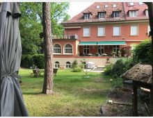 Rückansicht eines großen Wohnhauses mit rotem Putz und rotem Ziegeldach, mehreren Dachgauben und weißen Fensterläden. Im Vordergrund ein Garten mit Rasen, Bäumen, einem Sonnenschirm, Gartenmöbeln und Bepflanzung, umgeben von dichter Vegetation.