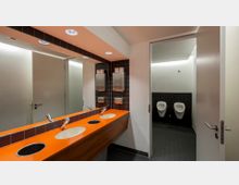 Eine moderne öffentliche Herrentoilette mit orangefarbener Waschfläche und eingelassenen Waschbecken sowie Edelstahlarmaturen, dahinter ein großer Spiegel und Wandspender. Rechts im Bild befinden sich zwei Urinale an einer mit schwarzen Fliesen verkleideten Wand.