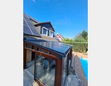 Ein modernes Einfamilienhaus mit dunkler Schindelfassade, Solarpaneelen auf dem Dach und einem angeschlossenen Wintergarten mit Glas- und Holzrahmenkonstruktion. Im Vordergrund ein Pool mit Holzterrasse, umgeben von Hecken und einem weiteren Gebäude im Hintergrund.
