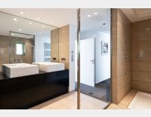 Modernes Badezimmer mit zwei rechteckigen weißen Waschbecken auf einem dunklen Waschtisch, großen beigen Wandfliesen, einer bodentiefen Dusche mit Glasabtrennung und einem beleuchteten Spiegel. Im Hintergrund ist ein offener Flur mit weißer Tür und Wanddekoration zu sehen.