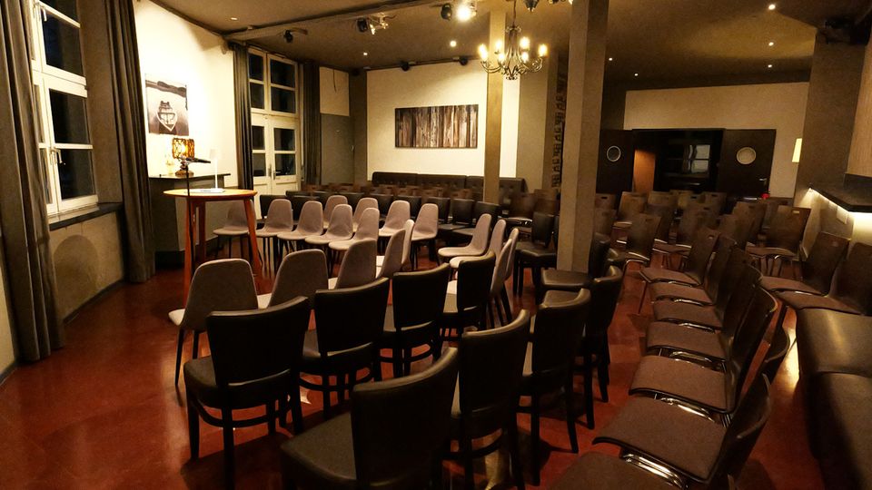 Innenansicht eines kleinen Veranstaltungs- oder Vortragsraums mit vielen in Reihen aufgestellten Stühlen, einer kleinen Bühne mit Rednerpult und großen Fenstern mit Vorhängen an der linken Wand. Der Raum hat eine hohe Decke mit Spotbeleuchtung und Kronleuchter, Säulen, dunkle Sitzbänke an den Wänden und einen rötlichen Boden.