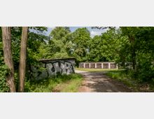 Eine mit Bäumen und Sträuchern bewachsene Landschaft mit einem unbefestigten Weg, der an einer Wand mit Graffiti vorbeiführt. Im Hintergrund stehen mehrere Garagen mit bemalten Rolltoren, umgeben von dichtem Grün.