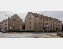 Mehrfamilienhäuser mit beigem Putz und roten Fensterrahmen an einer Straßenecke, umgeben von Grünflächen und Gehwegen. Verkehrsschilder zeigen eine Geschwindigkeitsbegrenzung von 50 km/h und eine Zone mit Tempo 30 an.