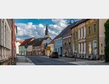 Straße in einer kleinen Stadt mit historischen Gebäuden und Häuserfassaden in verschiedenen Farben, darunter Fachwerk und Putzfassaden, sowie einem Kirchturm mit Uhr im Hintergrund. Links und rechts der Straße stehen Straßenlaternen und teilweise geparkte Autos, während der Himmel von weißen Wolken bedeckt ist.
