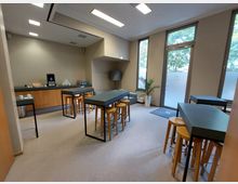 Moderner Aufenthaltsraum mit hohen Tischen und Hockern aus Holz, einer Kaffeestation auf einer Holztheke im Hintergrund und großen Fenstern, die natürliches Licht hereinlassen. Eine Glastür führt nach draußen, und ein Pflanzenkübel steht in der Ecke.