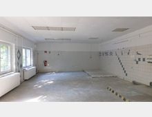 Ein leerer, rechteckiger Raum mit gefliesten Wänden und gefliestem Boden. An den weißen Wänden befinden sich blaue geometrische Muster, sowie ein Feuerlöscher; die Decke enthält mehrere eingelassene Lampen.