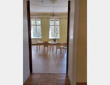 Blick in einen hellen Raum mit gelben Wänden und großen Fenstern, durch die Grünflächen sichtbar sind. Im Raum stehen Tische und Holzstühle auf einem hellen Boden, an der Decke hängt ein kleiner Kronleuchter.