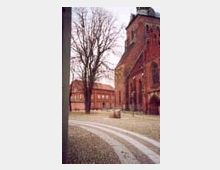 Backsteinkirche mit hohem Turm, umgeben von einer gepflasterten Fläche und kahlen Bäumen im Winter. Im Hintergrund ist ein weiteres rotes Backsteingebäude sichtbar.
