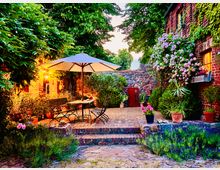 Gepflasterter Innenhof mit einem runden Metalltisch, umgeben von Gartenstühlen und einem großen Sonnenschirm. Die Umgebung ist von roten Backsteinmauern, blühenden Sträuchern, Topfpflanzen und einer Steinmauer geprägt, mit warmem Licht, das vom Haus auf die Szene scheint.