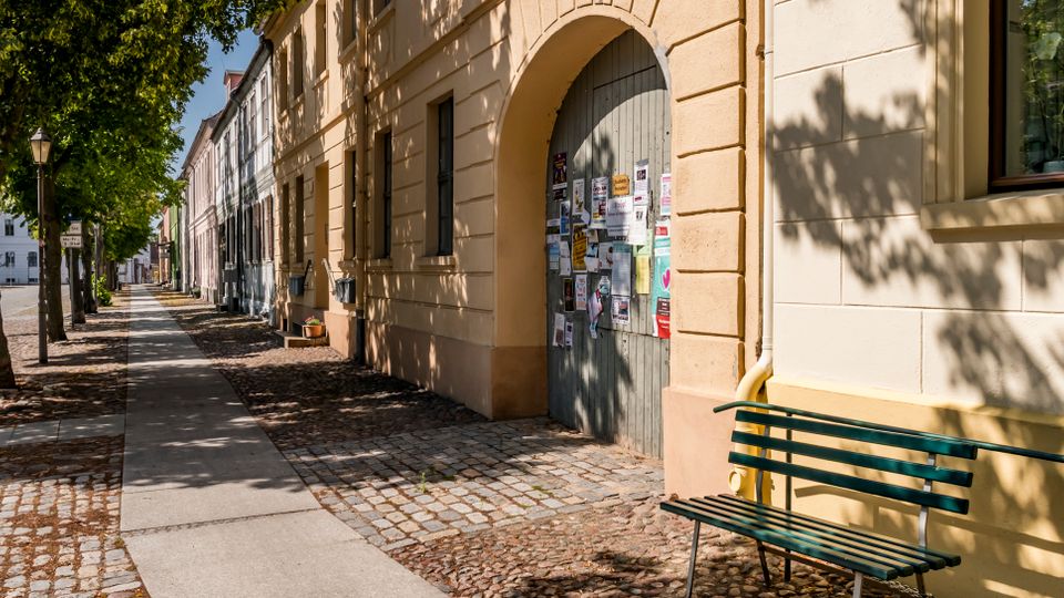 Gepflasterter Gehweg entlang einer von Bäumen gesäumten Straße mit historischen, hell verputzten Fassaden. Im Vordergrund stehen eine grüne Sitzbank und ein großes Torbogenportal mit angeschlagenen Plakaten.