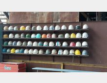 Regale mit zahlreichen bunten Schutzhelmen sind an einer rostfarbenen Metallwand angebracht, darunter ein roter Behälter mit der Aufschrift „Sand“. Die Umgebung wirkt industriell, mit sichtbaren Stahlkonstruktionen im Hintergrund.