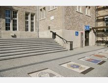 Außenansicht eines Gebäudes aus Naturstein mit großen rechteckigen Fenstern, einer breiten Treppe und einer barrierefreien Eingangstür mit Rollstuhlsymbol. Auf dem Pflasterboden davor befinden sich Mosaikbilder mit verschiedenen Motiven.