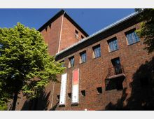 Backstein-Gebäude mit großen, rechteckigen Fenstern und einem Turm, umgeben von Bäumen mit grünem Laub vor einem klaren blauen Himmel. An der Fassade hängen zwei vertikale Banner mit Schriftzügen.
