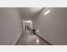 Langer, schlichter Flur eines Gebäudes mit weißen Wänden, mehreren Türen auf der linken Seite und grauen Sitzgelegenheiten entlang der rechten Wand. Der Boden ist hell, und kleine Wandlampen sorgen für Beleuchtung.