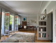 Wohnzimmer mit Holzboden, großen französischen Fenstern und Blick auf einen Balkon mit grüner Gartenlandschaft. Der Raum ist mit einem großen Teppich, einem Esstisch, Stühlen, einem antiken Holzschrank und Wanddekorationen ausgestattet.