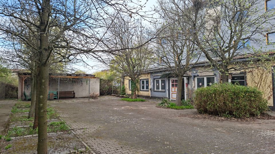 Innenhof mit gepflasterter Fläche und mehreren kahlen Bäumen, umgeben von niedrigen Gebäudeteilen mit Fenstern und einer Tür mit farbigem Rahmen. Links steht ein offener Unterstand mit Sitzbank, rechts sind Sträucher und kleine Grünflächen am Rand zu sehen.