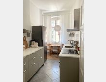 Schmale Küche mit hellen Wänden und grauen Bodenfliesen, ausgestattet mit einer modernen grauen Küchenzeile, integriertem Herd und Spüle. Am Ende des Raums steht ein runder Holztisch mit vier Stühlen vor einem großen Fenster mit weißen Vorhängen, ergänzt durch eine hängende Papierlampe.