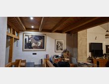 Wohnraum mit Holzbalkendecke und beleuchteten Spotlights, weißen Wänden und Wandkunst, darunter ein großes Gemälde und ein gerahmtes Foto. Im Raum stehen Bücherregale, ein Fernseher, Möbelstücke und verschiedene Dekorationsgegenstände.