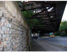 Backsteinmauer entlang eines Gehwegs unter einer rostigen Stahlbrücke, mit umliegender Vegetation und gepflasterter Straße im Hintergrund. Zwei Personen sind auf dem Weg nahe der Brücke zu erkennen.