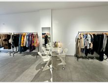 Ein modernes Bekleidungsgeschäft mit schlichten weißen Wänden und grauem Fliesenboden. Kleiderstangen mit farbenfroher und neutraler Kleidung stehen entlang der Wände, in der Mitte befinden sich ein kleiner weißer Tisch und zwei moderne weiße Stühle.