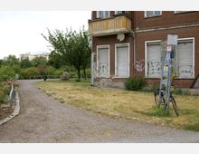 Eine Grünfläche mit einem Kiesweg, Rasen und Büschen; im Vordergrund ein Eck eines braunen Wohnhauses mit heruntergelassenen Rolläden und Graffiti. Im Hintergrund sind weitere Bäume und ein mehrstöckiges Wohnhaus erkennbar.