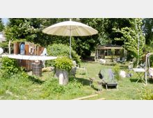 Grüne Gartenlandschaft mit Rasenfläche, Sonnenschirm und verschiedenen Gartenmöbeln. Im Hintergrund steht eine kleine Holzhütte mit Sitzgelegenheiten, umgeben von Bäumen und Büschen.