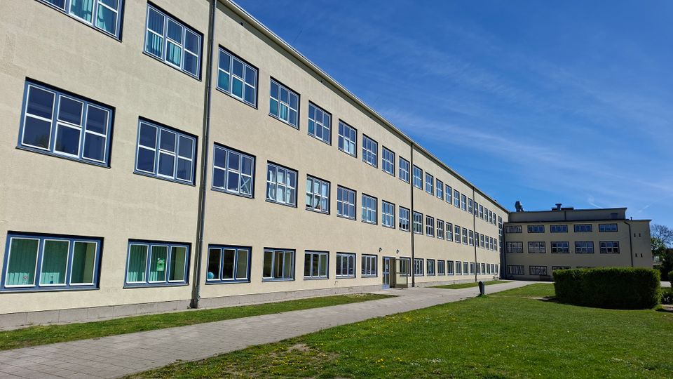 Langes, mehrstöckiges Gebäudetrakt mit heller Putzfassade und Reihen gleichförmiger Fenster, aufgenommen aus schräger Perspektive. Davor verlaufen ein gepflasterter Weg und eine Grünfläche; im Hintergrund ist ein weiterer Gebäudeflügel unter blauem Himmel zu sehen.
