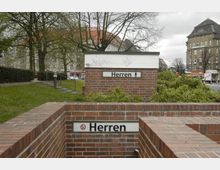 Backsteineingang zu einer öffentlichen Herrentoilette in einer Stadtumgebung, mit Beschilderung "Herren" und umliegendem Grünbereich sowie mehrstöckigen Gebäuden im Hintergrund. Im oberen Bereich ist eine Wand mit Graffiti zu sehen, und es befinden sich weitere Verkehrs- und Geschäftsanzeigen im Hintergrund.