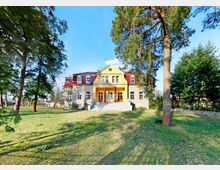 Historisches Gebäude mit gelber Fassade und rotem Dach, umgeben von einer weitläufigen Grünfläche mit Bäumen und blühenden Pflanzen im Vordergrund; breite Außentreppe führt zum Haupteingang mit Säulen.