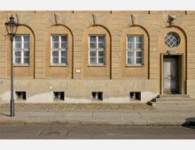 Fassade eines historischen Gebäudes mit beigem Verputz, mehreren hohen Fenstern mit Bögen, einem verzierten Holzeingang mit Stufen und einem ovalen Fenster darüber. Links steht eine alte Straßenlaterne auf dem Gehweg aus Kopfsteinpflaster und Betonplatten.