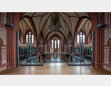 Innenansicht einer modernen Veranstaltungshalle in einer neugotischen Kirche. Zu sehen sind hohe Spitzbögen, rote Backsteinwände, schmale Fenster mit gotischem Stil sowie moderne Einbauten wie Metallgeländer, Glasflächen und Zwischengalerien.